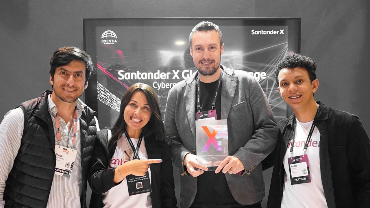 A imagem mostra um grupo de pessoas posando para uma foto durante a cerimônia de premiação do Santander X Global Challenge. Existem quatro indivíduos sorrindo. Rafael Umann, CEO da Azion, está no centro, segurando o prêmio.